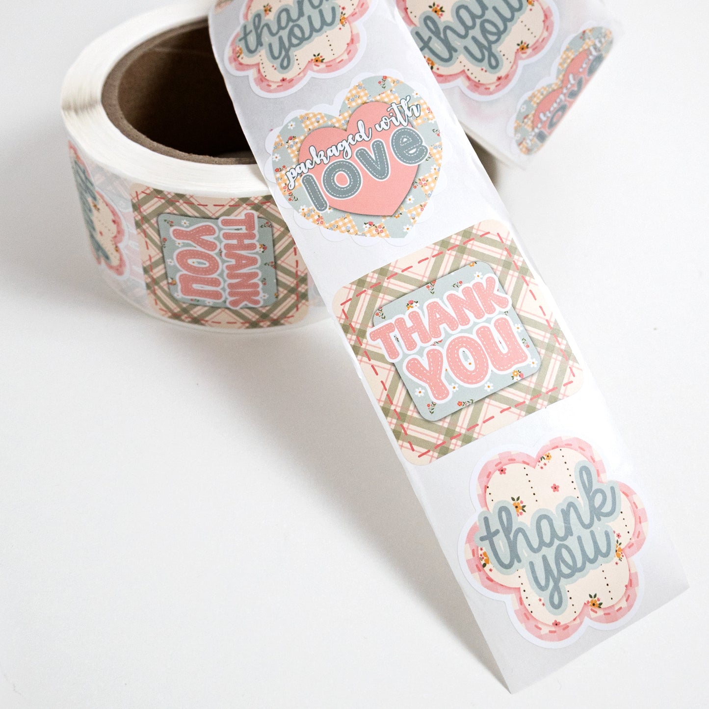 A roll of cute, die cut, small business packaging stickers in a country cottage theme with a repeating pattern of 'thank you' and 'packaged with love' labels in pink, powder blue, beige, and olive plaid, shown on a clean white background.