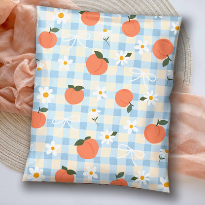 A 10x13 poly mailer with a light blue and cream gingham pattern, decorated with peaches illustrations, white daisies, and white bows. The shipping bag is styled on a woven mat with soft peach fabric accents.