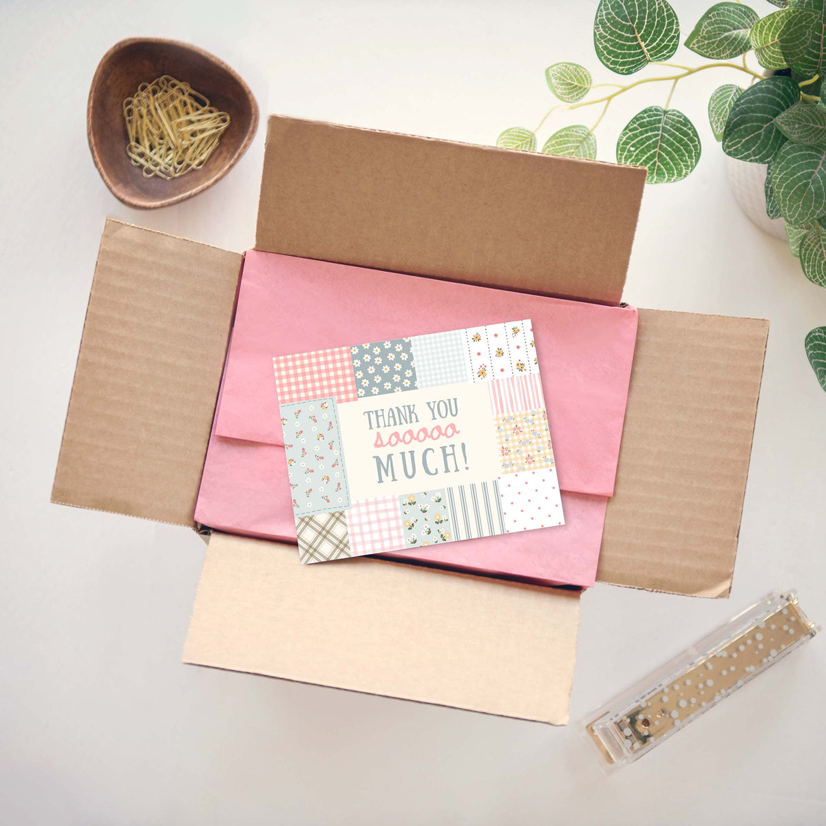 A flat-lay view of a customer thank you message card placed inside an open brown cardboard box lined with pink tissue paper. The card features a colorful patchwork pattern of gingham, stripes, and polka dots with the text 'THANK YOU sooooo MUCH!'.