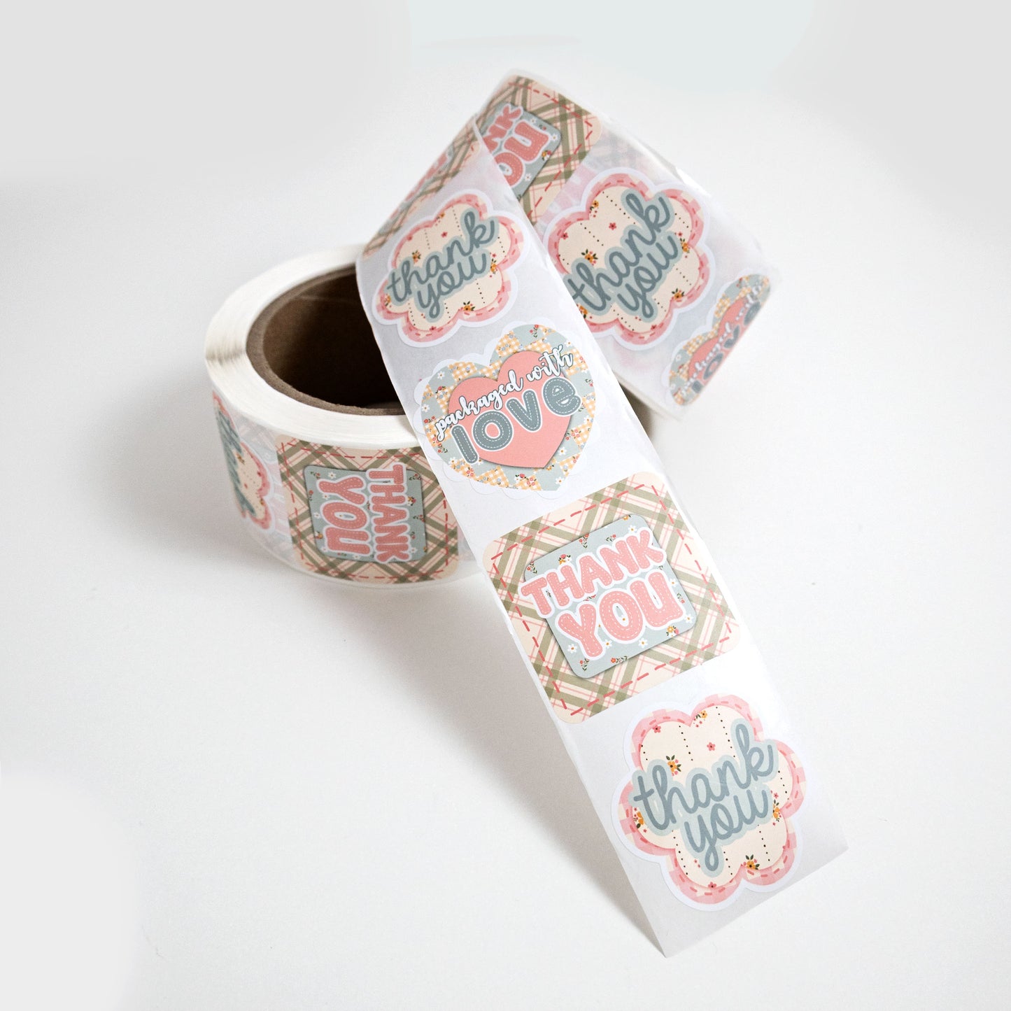 A roll of cute, die cut, small business packaging stickers in a country cottage theme with a repeating pattern of 'thank you' and 'packaged with love' labels in pink, powder blue, and beige plaid, shown on a clean white background.