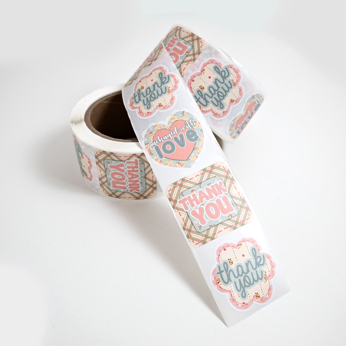 A roll of cute, die cut, small business packaging stickers in a country cottage theme with a repeating pattern of 'thank you' and 'packaged with love' labels in pink, powder blue, and beige plaid, shown on a clean white background.