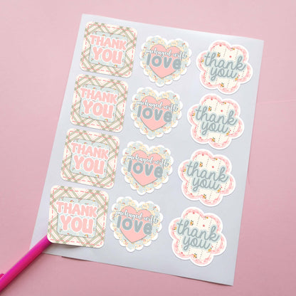 A sheet of die-cut customer thank you stickers. The stickers feature phrases like 'thank you' and 'packaged with love' in soft pastel colors, including pink, beige, blue, and white, with plaid, floral, and scalloped patchwork designs. For small business or boutique customer packaging