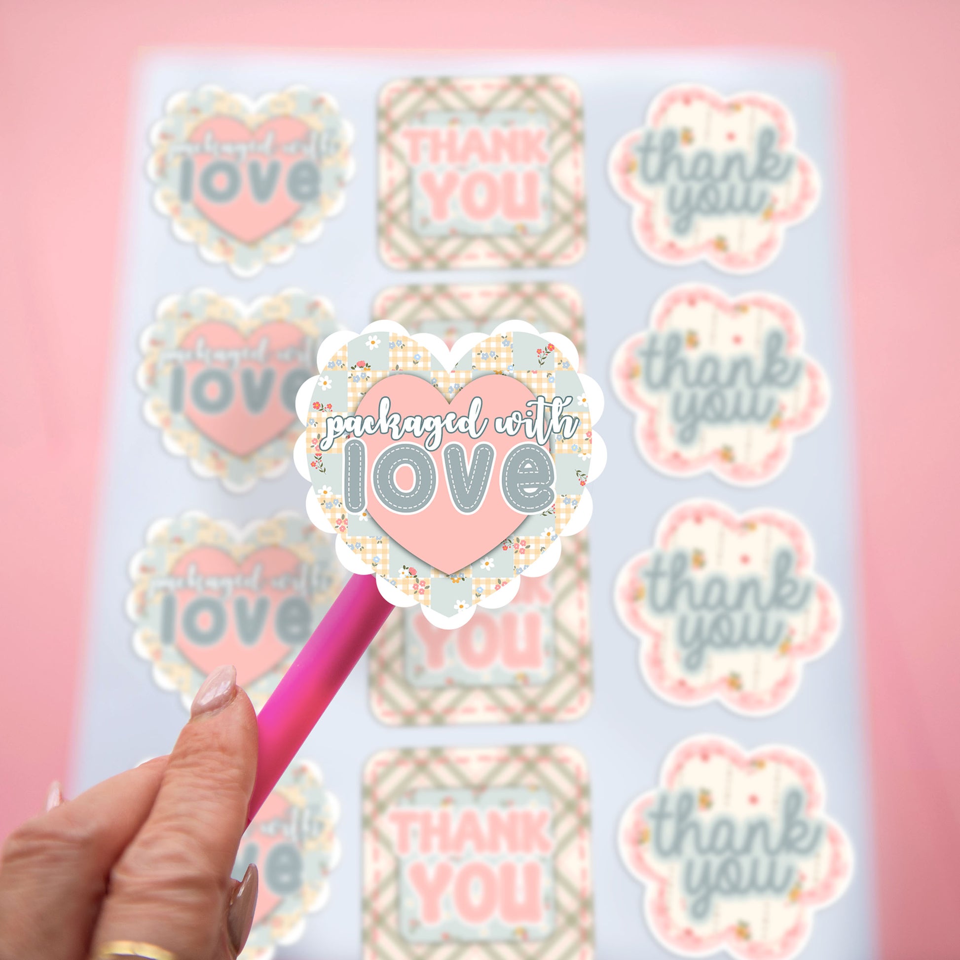 A hand holds a pink pen-like tool with a heart-shaped sticker in front of a sheet of matching cusotmer packaging stickers. The stickers read phrases like 'thank you' and 'packaged with love' in pastel pink, blue-gray, and cream tones with small patchwork details, against a soft pink background.