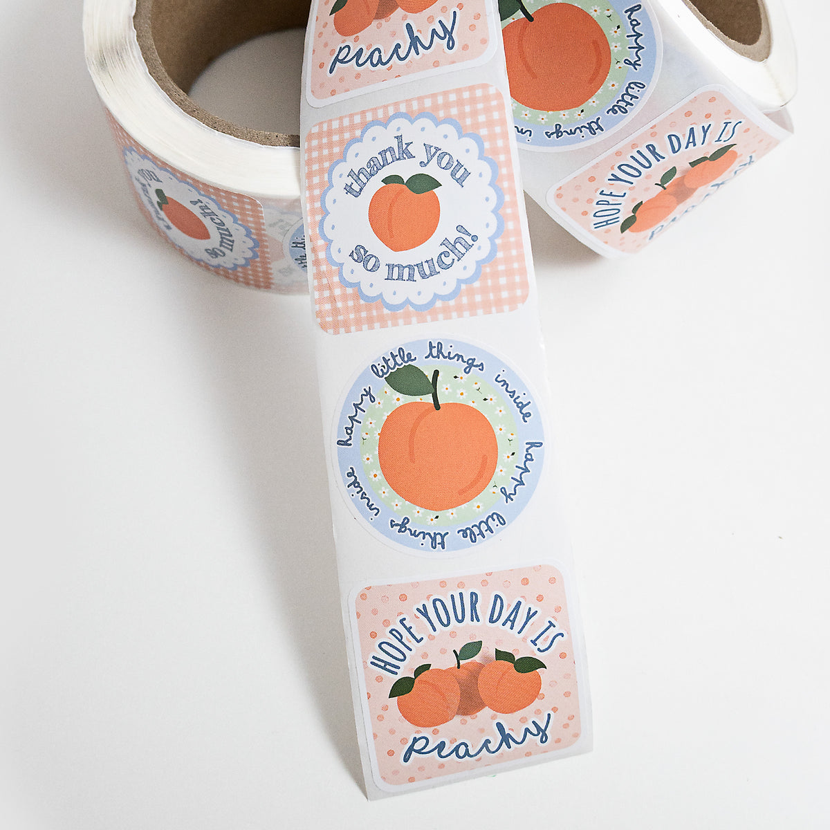 A roll of die-cut customer packaging thank you stickers with peach-themed labels, shown partially unrolled on a white background. The labels feature illustrations of peaches, gingham patterns, and handwritten text such as 'thank you so much!' and 'hope your day is peachy'